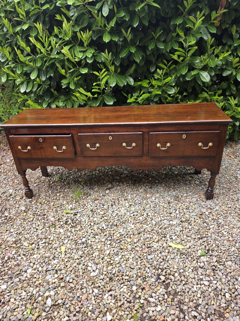 A mid-18th-century Georgian oak dresser base featuring three frieze drawers with brass swan-neck handles, a shaped apron, and turned front legs. View VII