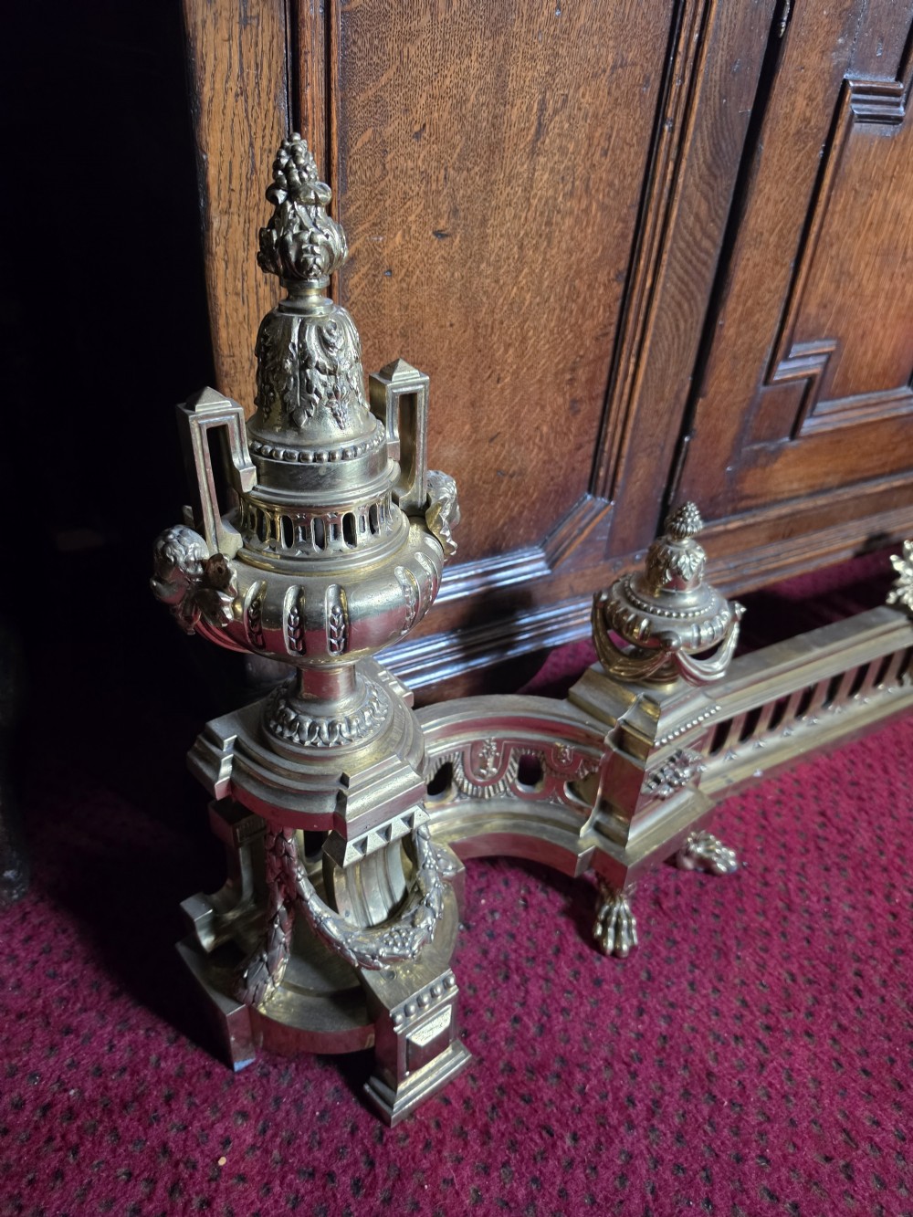 An ornate Louis XVI style brass fireplace fender featuring urn finials, classical mask details, and a pierced decorative rail. View II