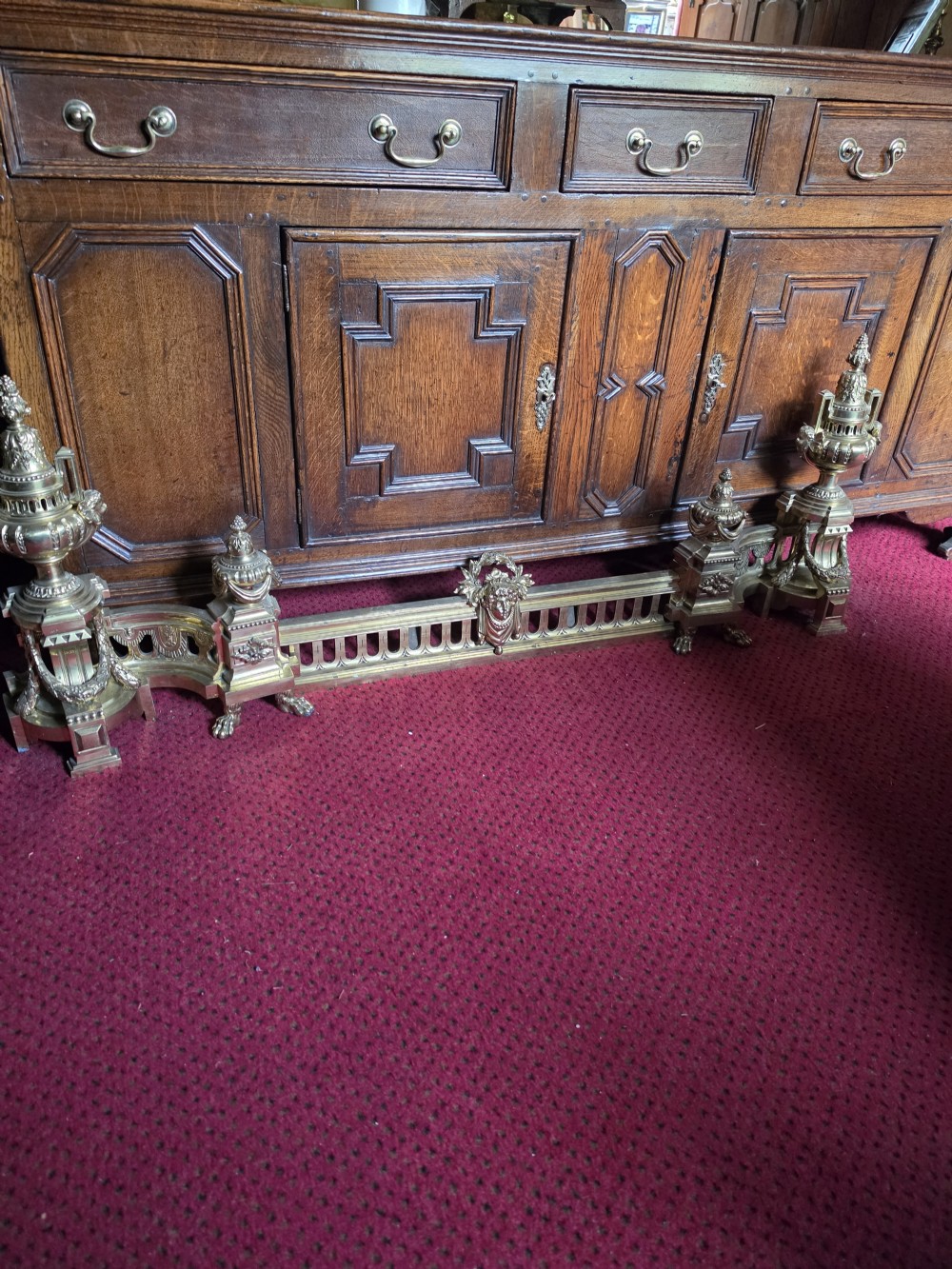 An ornate Louis XVI style brass fireplace fender featuring urn finials, classical mask details, and a pierced decorative rail.