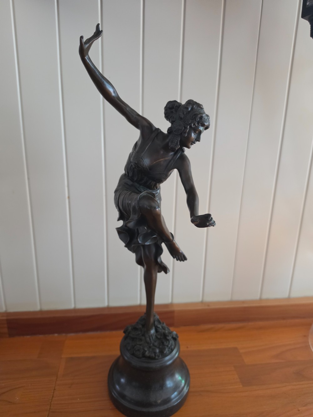 A dark patinated bronze figure depicting a female dancer in mid-motion with raised arms, standing balanced on a circular base.