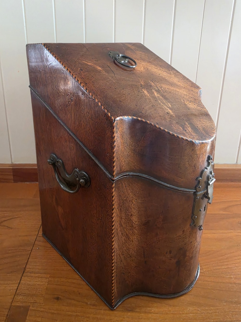 A Georgian mahogany knife box featuring a sloped lid, serpentine front, brass lock plate, ring handle, and edge stringing. View V