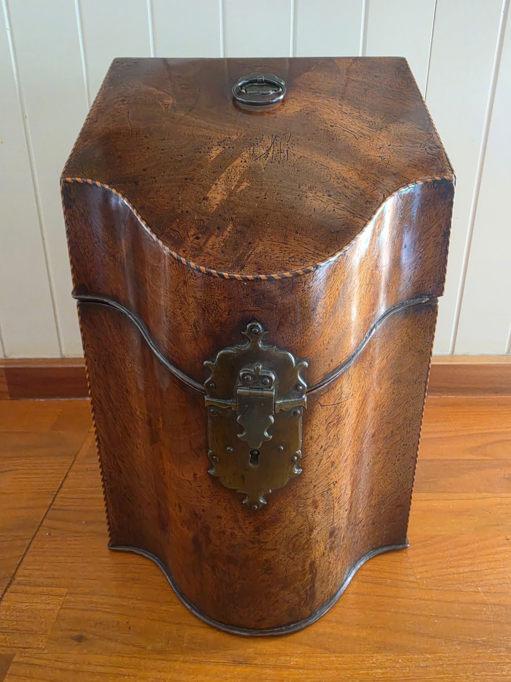 A Georgian mahogany knife box featuring a sloped lid, serpentine front, brass lock plate, ring handle, and edge stringing. View VI