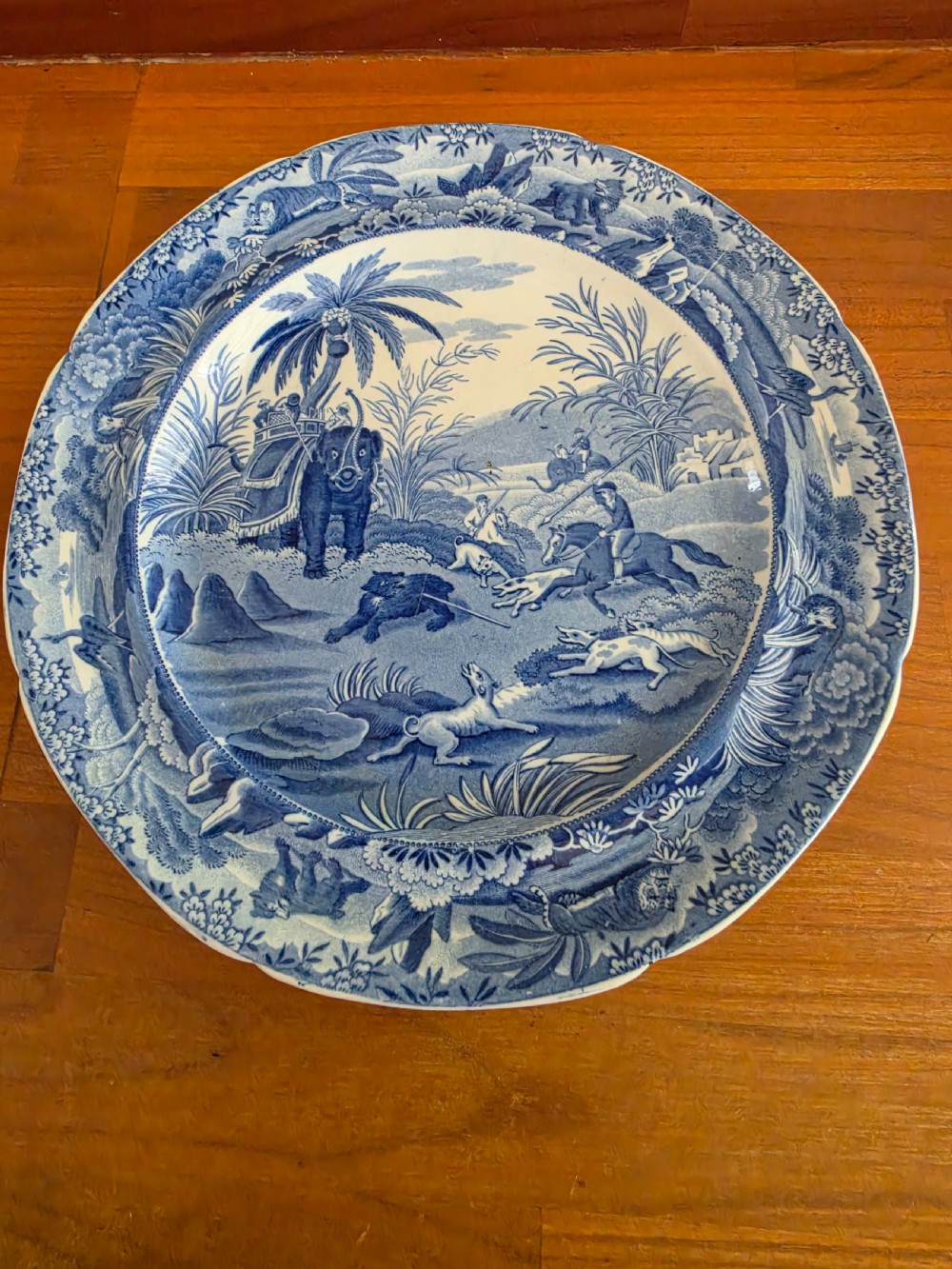A circular blue and white ceramic transferware plate depicting a classical Indian hunting scene with horses, hounds, an elephant, and a bear.