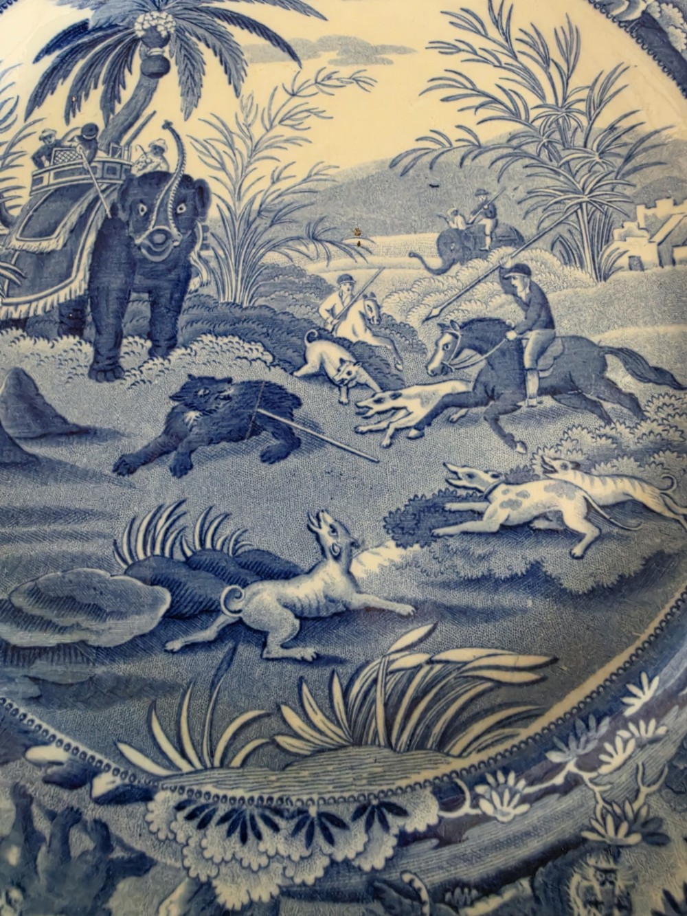 A circular blue and white ceramic transferware plate depicting a classical Indian hunting scene with horses, hounds, an elephant, and a bear. View II