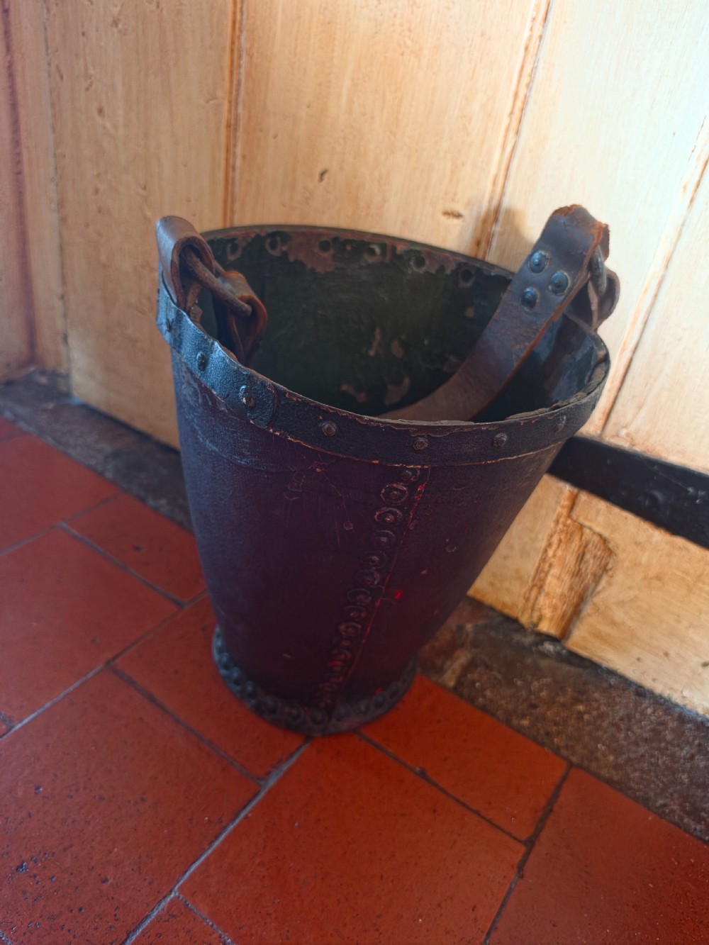 A conical dark brown leather fire bucket with metal studding along the rim and side seams and an attached leather handle.