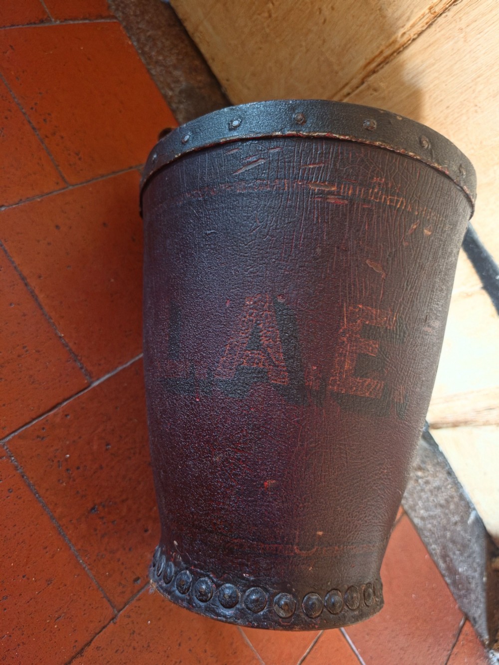 A conical dark brown leather fire bucket with metal studding along the rim and side seams and an attached leather handle. View II