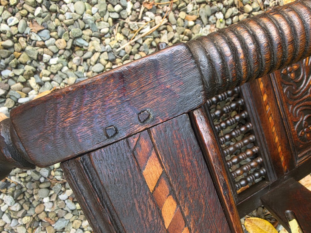 A 17th-century dark oak wainscot armchair featuring a carved back panel with floral inlay, turned front legs, scrolled cresting, and wooden peg joinery. View IX