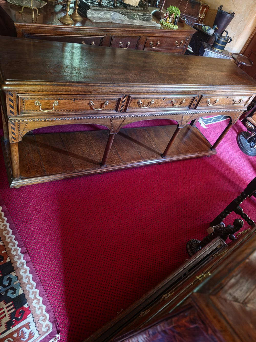 A 19th-century dark oak potboard dresser base featuring three inline drawers with brass swan-neck handles, geometric inlay details, carved arches, and an open lower shelf. View IV