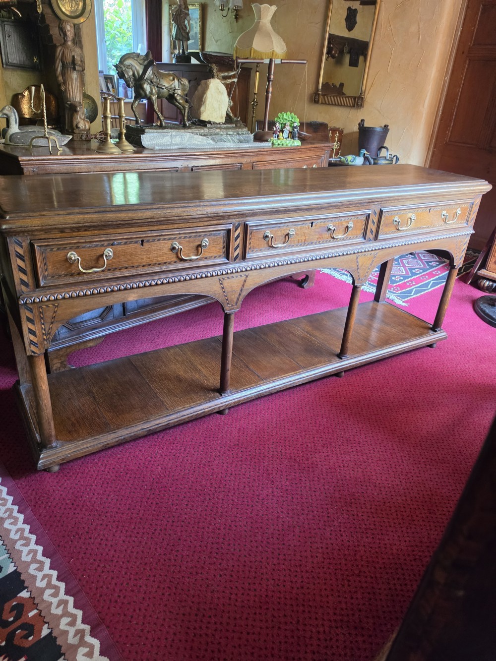 A 19th-century dark oak potboard dresser base featuring three inline drawers with brass swan-neck handles, geometric inlay details, carved arches, and an open lower shelf. View VI