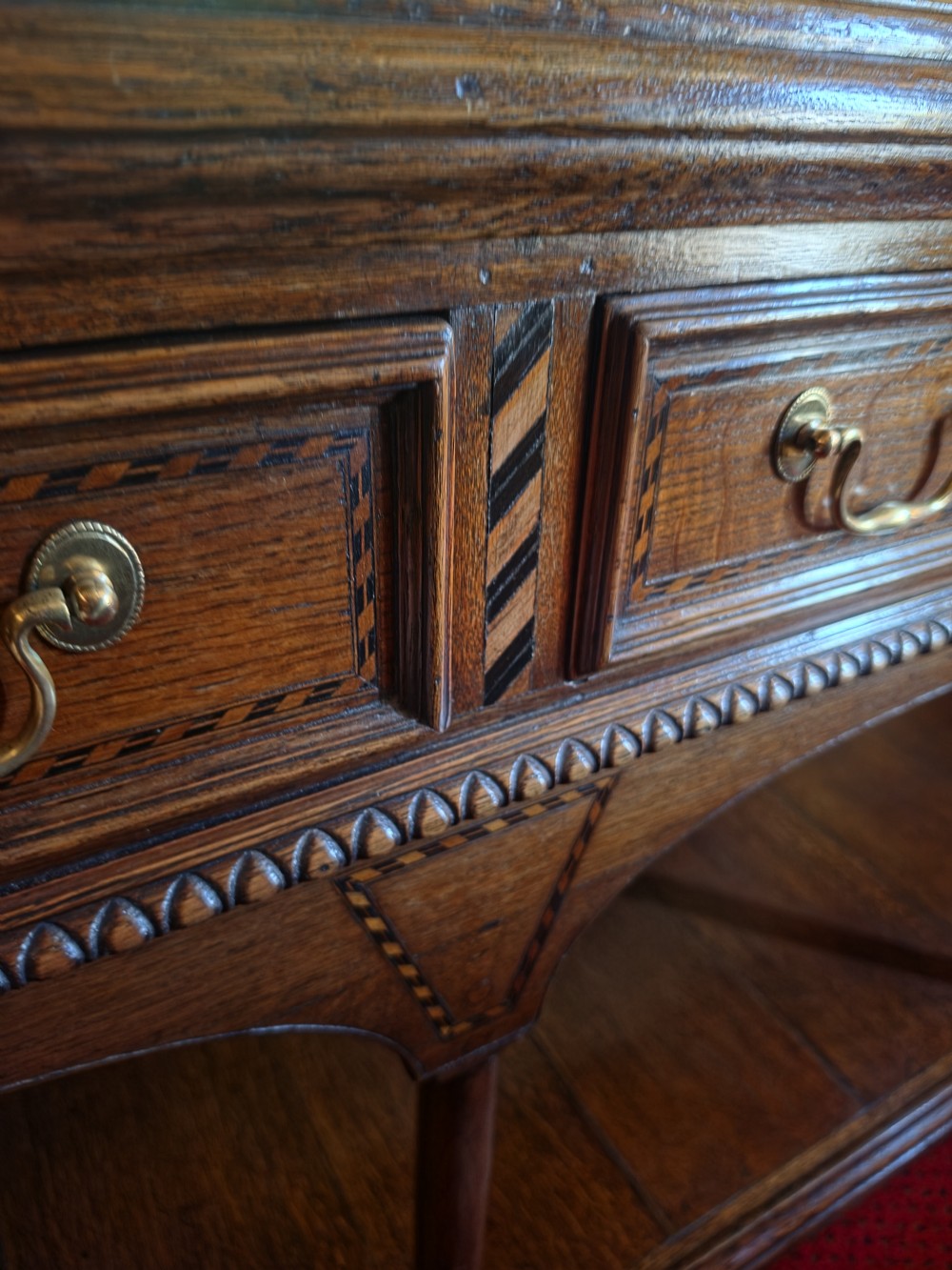 A 19th-century dark oak potboard dresser base featuring three inline drawers with brass swan-neck handles, geometric inlay details, carved arches, and an open lower shelf. View VII