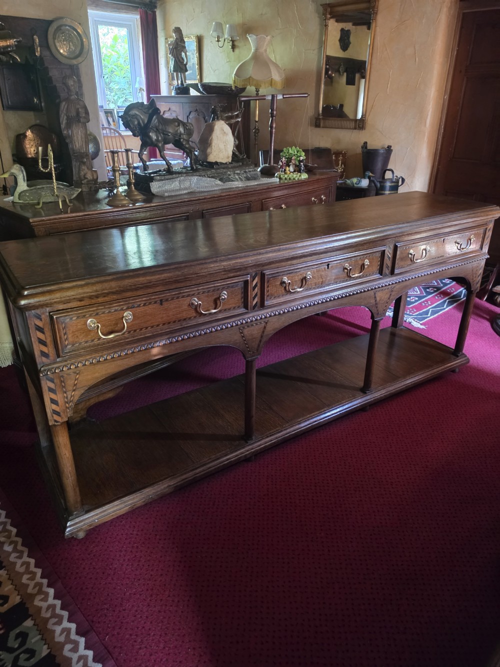 A 19th-century dark oak potboard dresser base featuring three inline drawers with brass swan-neck handles, geometric inlay details, carved arches, and an open lower shelf. View VIII