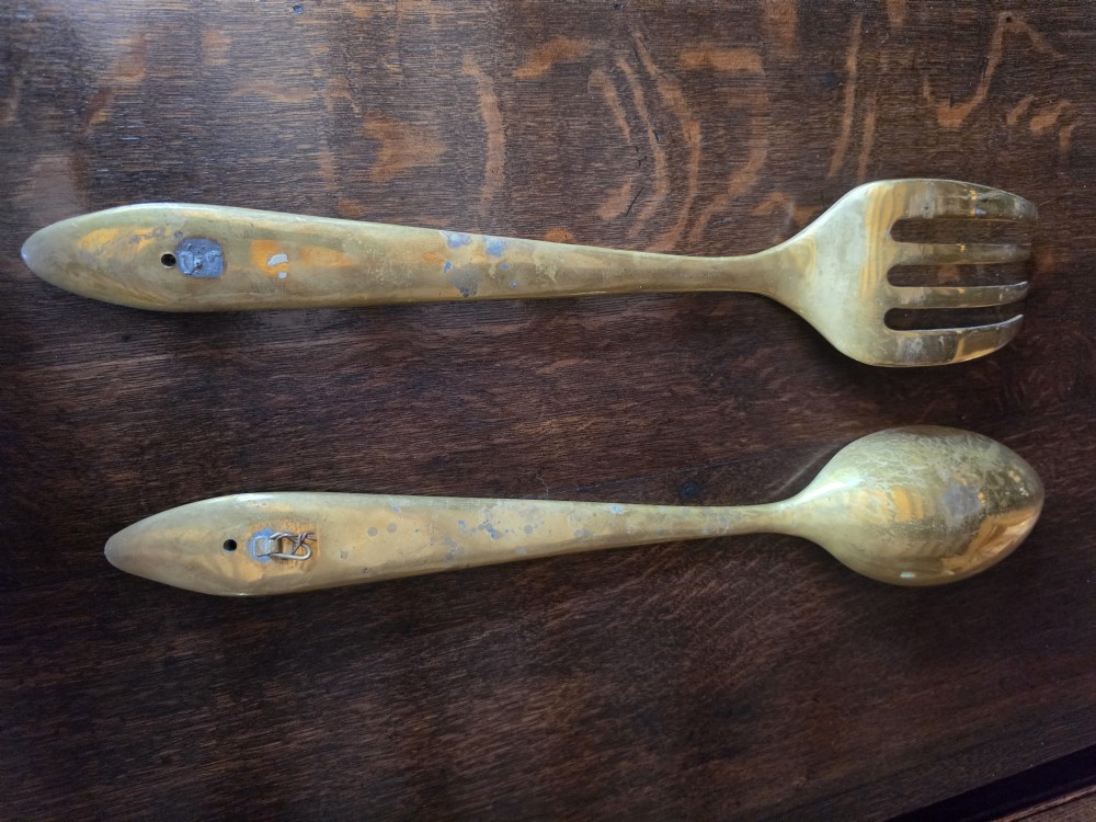 A pair of vintage brass wall plaques shaped as a large spoon and fork with pre-drilled mounting holes at the top of the handles. View II