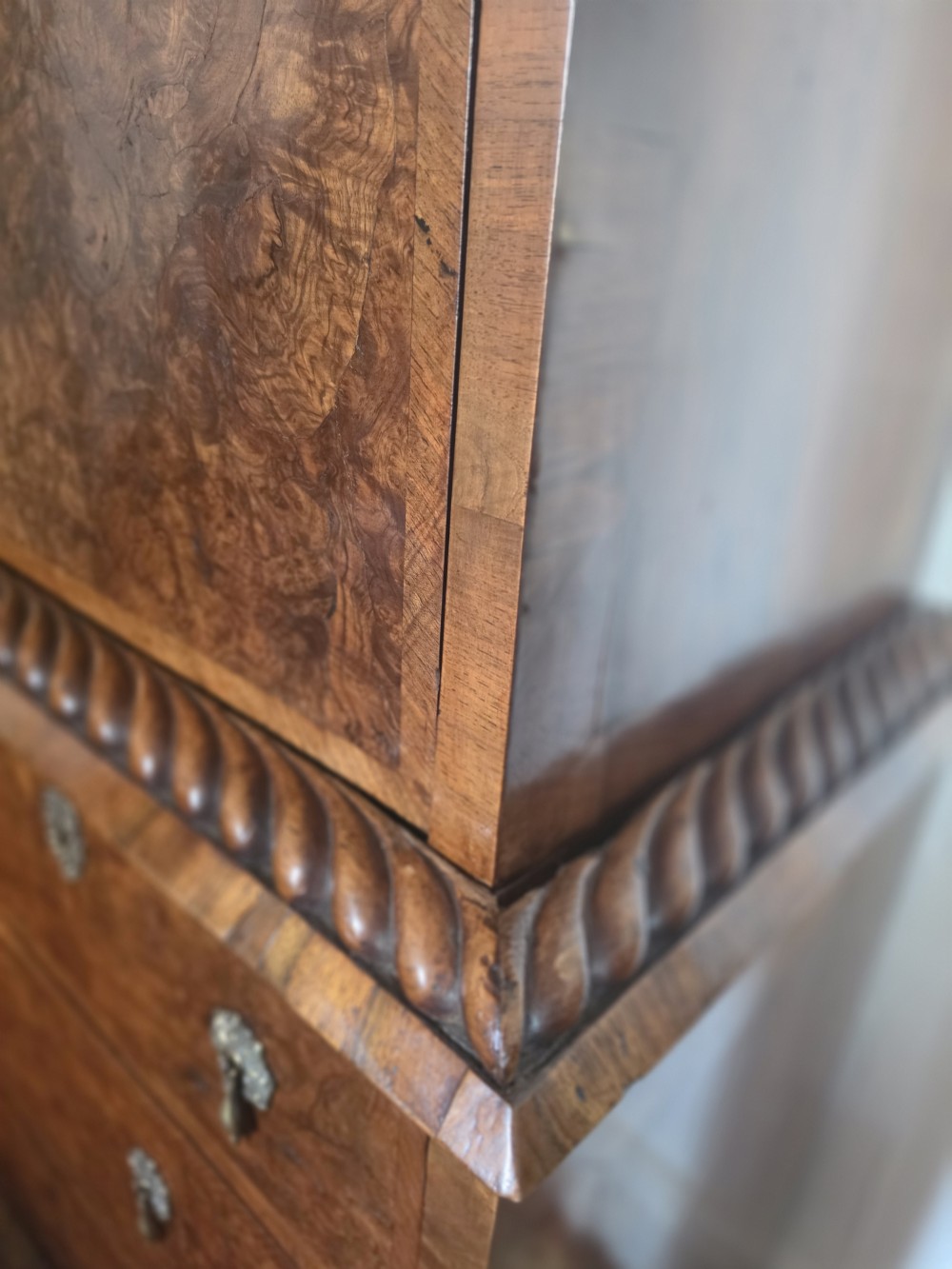 An early eighteenth-century English burr walnut escritoire featuring a carved architectural crest, fall-front writing surface, brass drop handles, and a chest of drawers base resting on bracket feet. View II