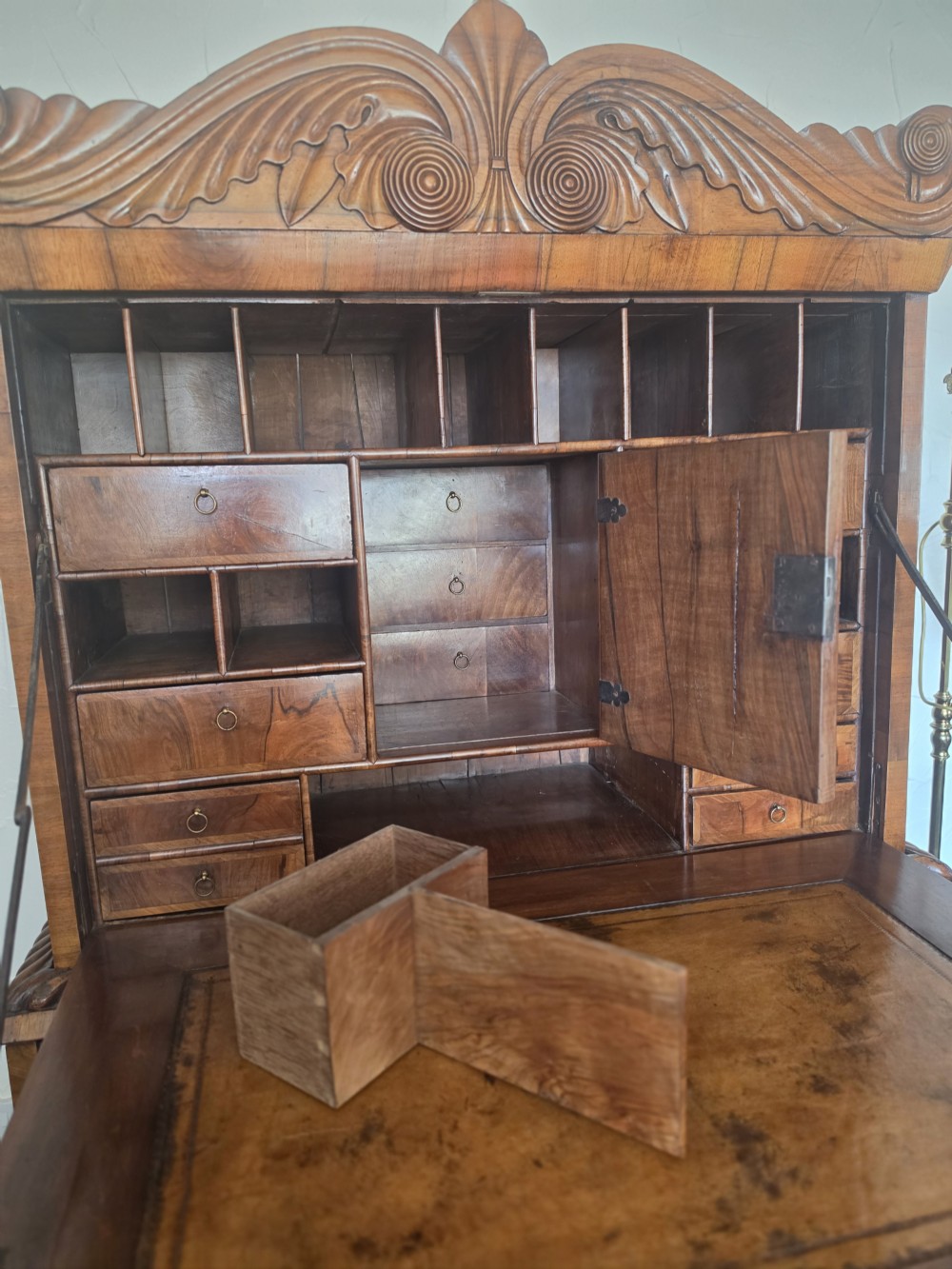 An early eighteenth-century English burr walnut escritoire featuring a carved architectural crest, fall-front writing surface, brass drop handles, and a chest of drawers base resting on bracket feet. View V