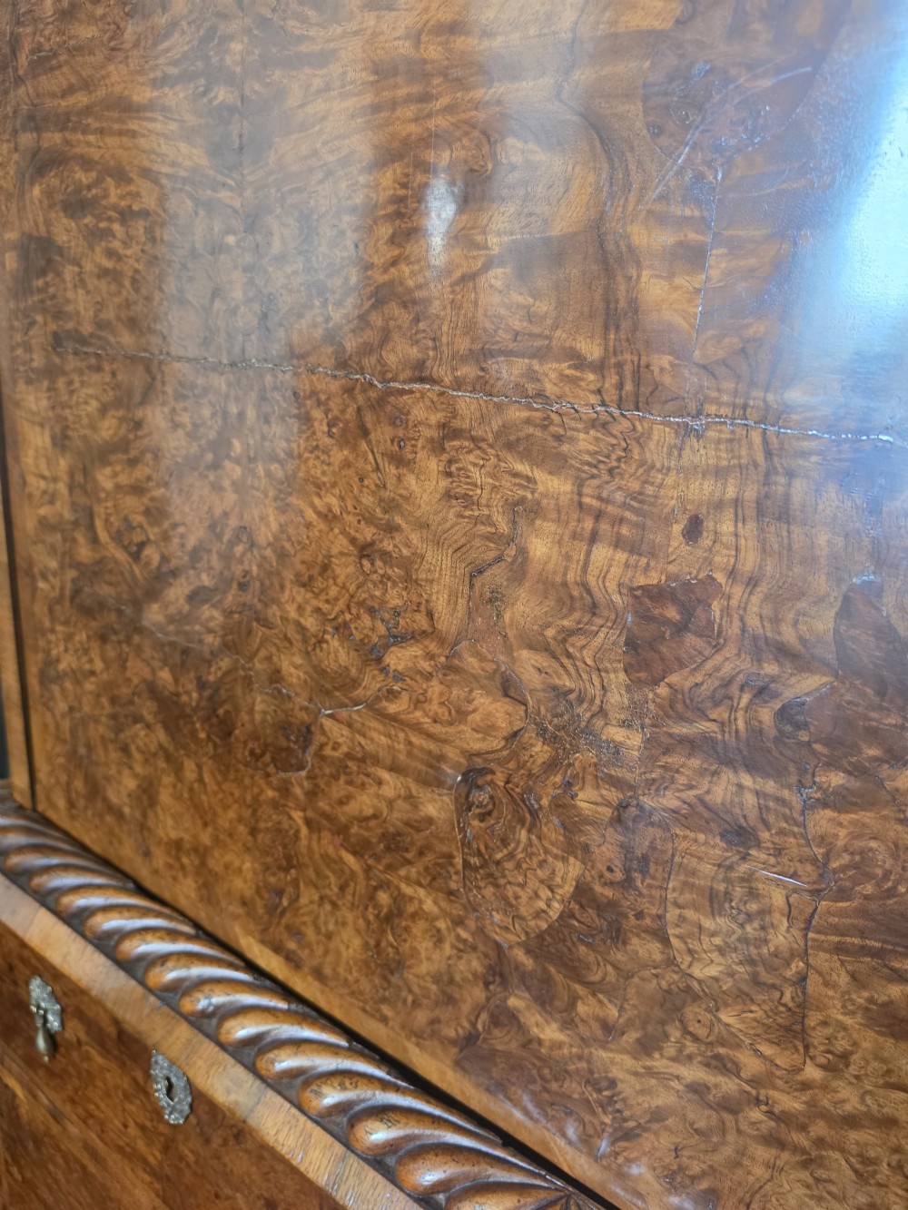 An early eighteenth-century English burr walnut escritoire featuring a carved architectural crest, fall-front writing surface, brass drop handles, and a chest of drawers base resting on bracket feet. View VI