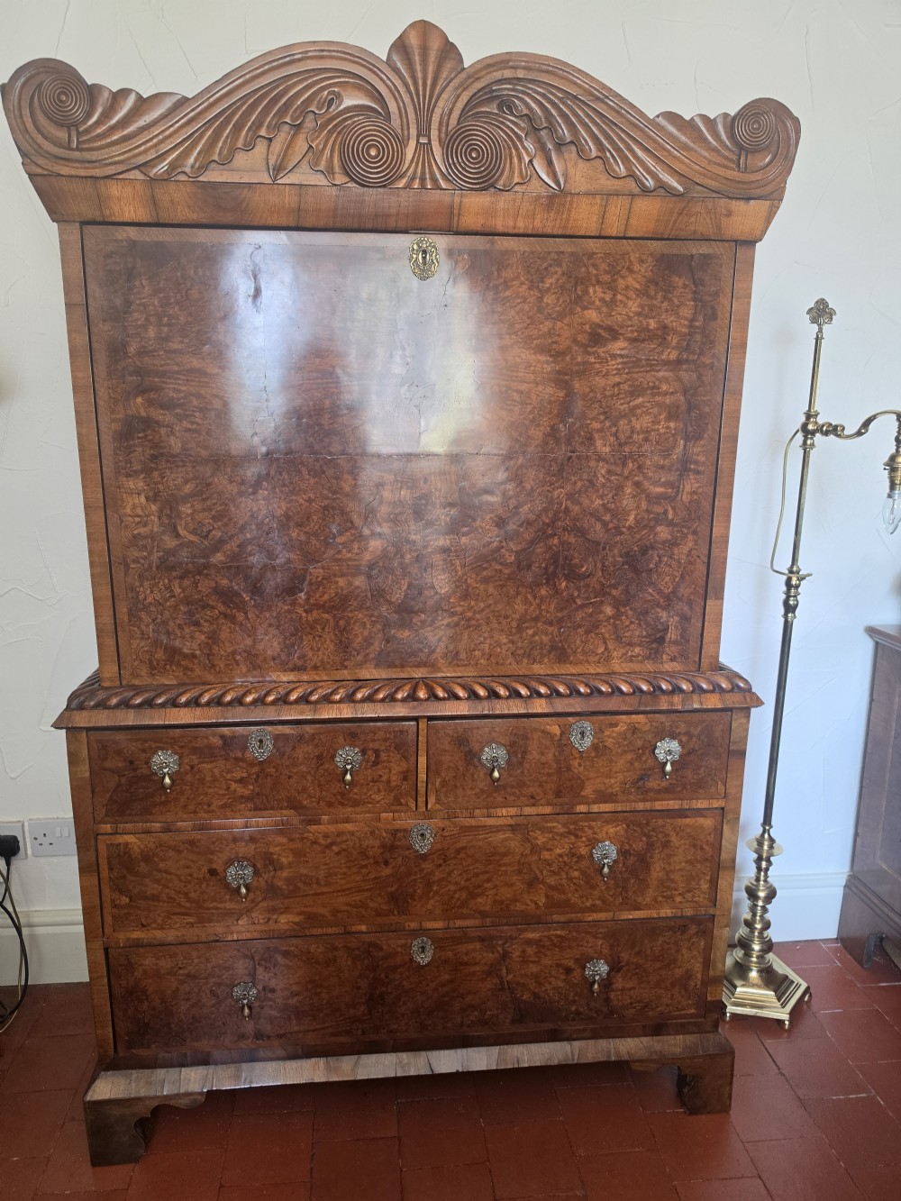 An early eighteenth-century English burr walnut escritoire featuring a carved architectural crest, fall-front writing surface, brass drop handles, and a chest of drawers base resting on bracket feet. View VII