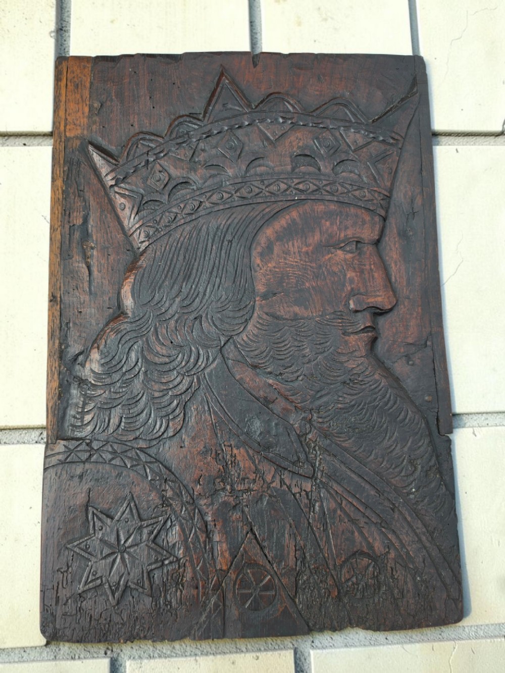 A rectangular dark oak carved panel featuring a profile relief portrait of a crowned king with a beard and long hair.