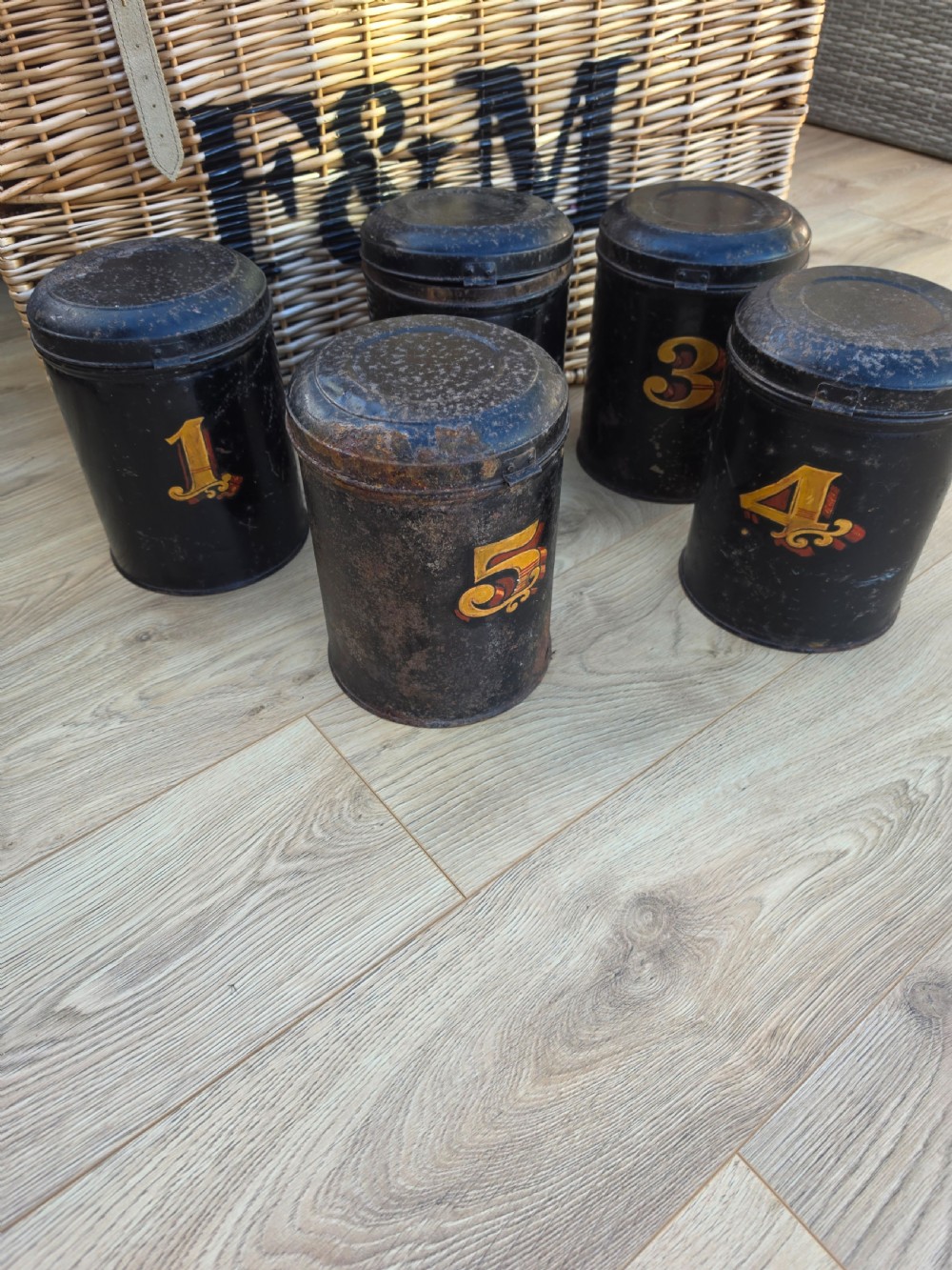 A set of five cylindrical black tin tea canisters numbered one through five with ornate gold and red painted numerals. View IV