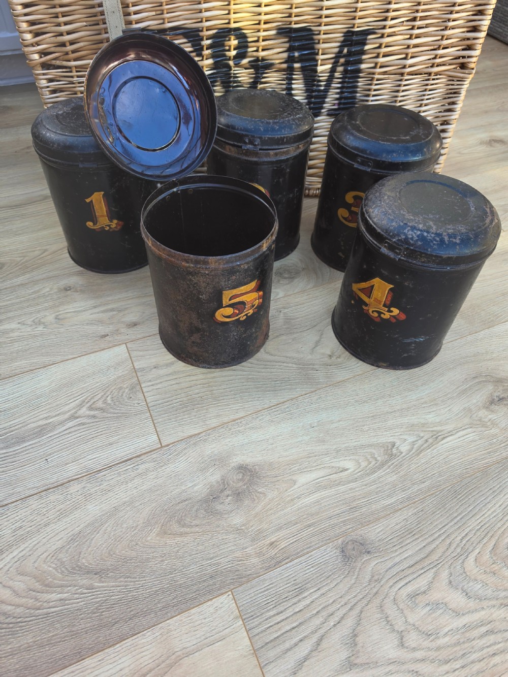 A set of five cylindrical black tin tea canisters numbered one through five with ornate gold and red painted numerals. View V