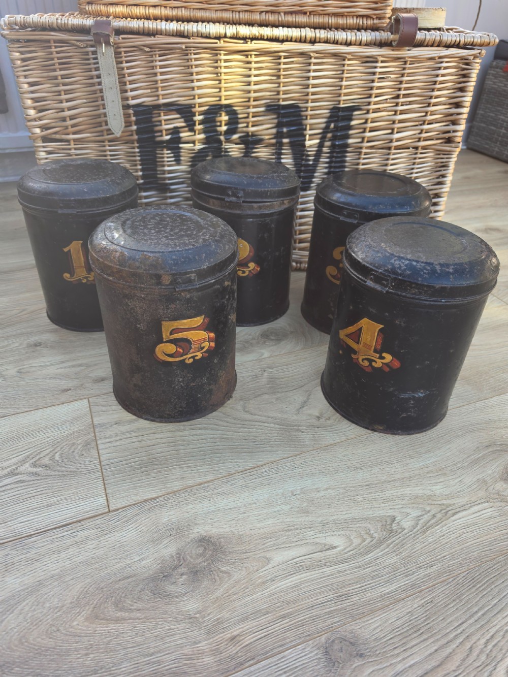 A set of five cylindrical black tin tea canisters numbered one through five with ornate gold and red painted numerals. View VI