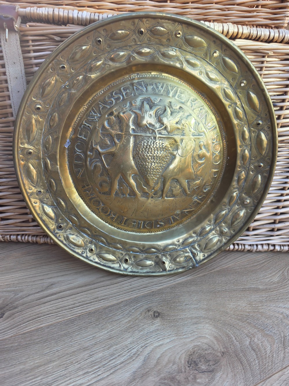 A circular 17th-century brass alms dish featuring central repousse decoration of two figures carrying a large cluster of grapes, surrounded by embossed inscriptions and decorative rim borders.
