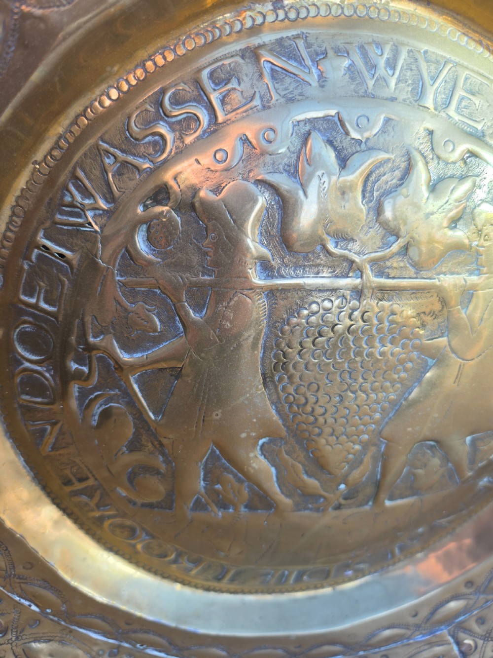 A circular 17th-century brass alms dish featuring central repousse decoration of two figures carrying a large cluster of grapes, surrounded by embossed inscriptions and decorative rim borders. View II