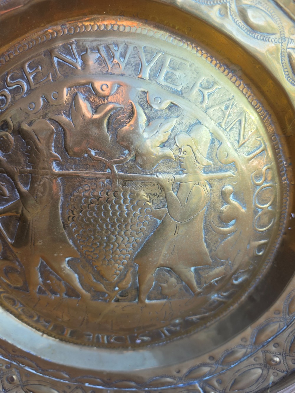 A circular 17th-century brass alms dish featuring central repousse decoration of two figures carrying a large cluster of grapes, surrounded by embossed inscriptions and decorative rim borders. View III