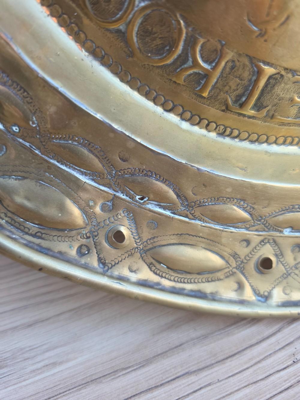 A circular 17th-century brass alms dish featuring central repousse decoration of two figures carrying a large cluster of grapes, surrounded by embossed inscriptions and decorative rim borders. View IV