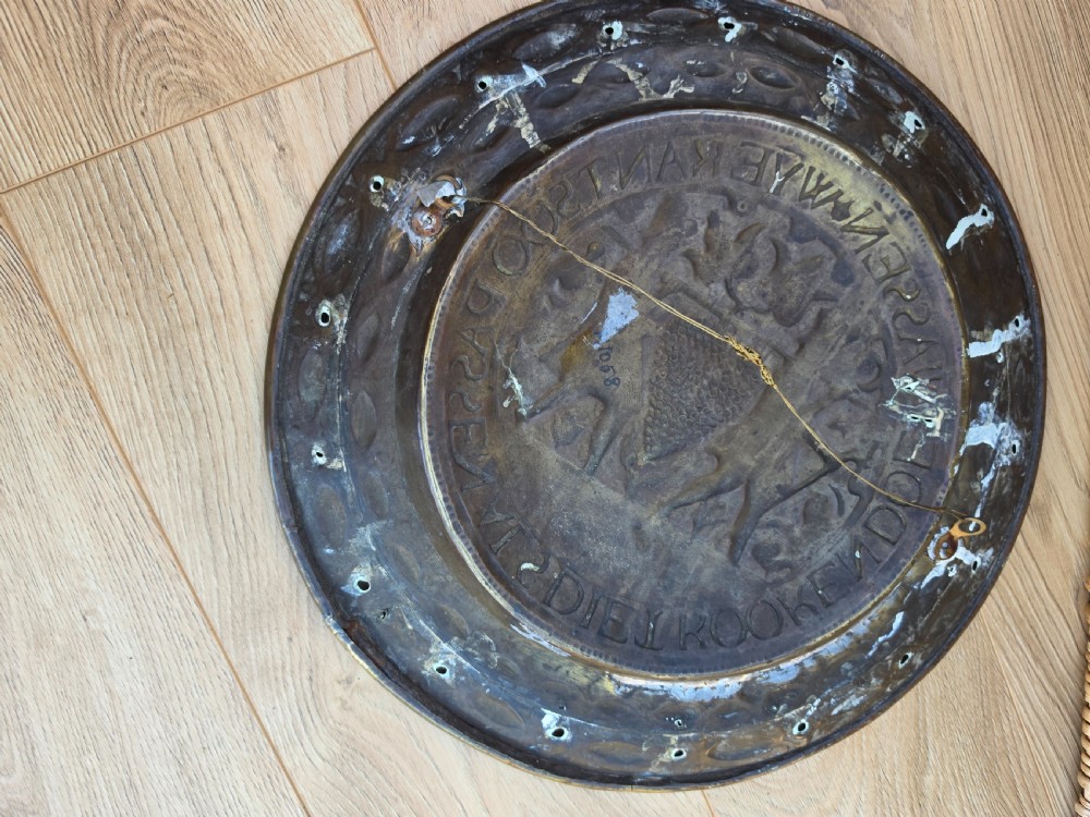 A circular 17th-century brass alms dish featuring central repousse decoration of two figures carrying a large cluster of grapes, surrounded by embossed inscriptions and decorative rim borders. View V
