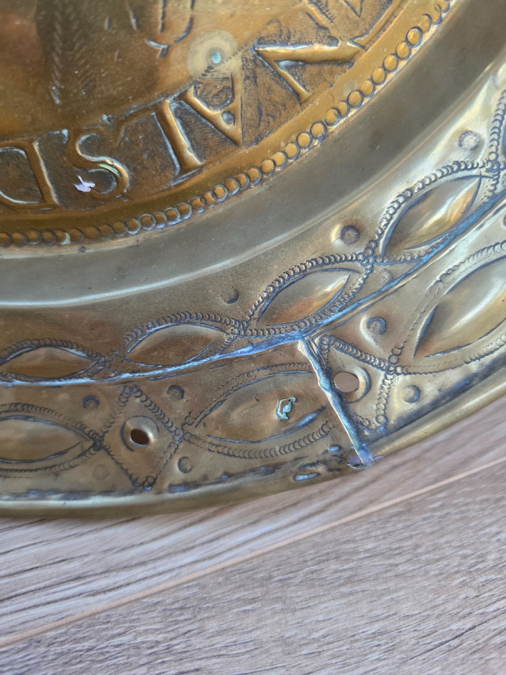 A circular 17th-century brass alms dish featuring central repousse decoration of two figures carrying a large cluster of grapes, surrounded by embossed inscriptions and decorative rim borders. View VI