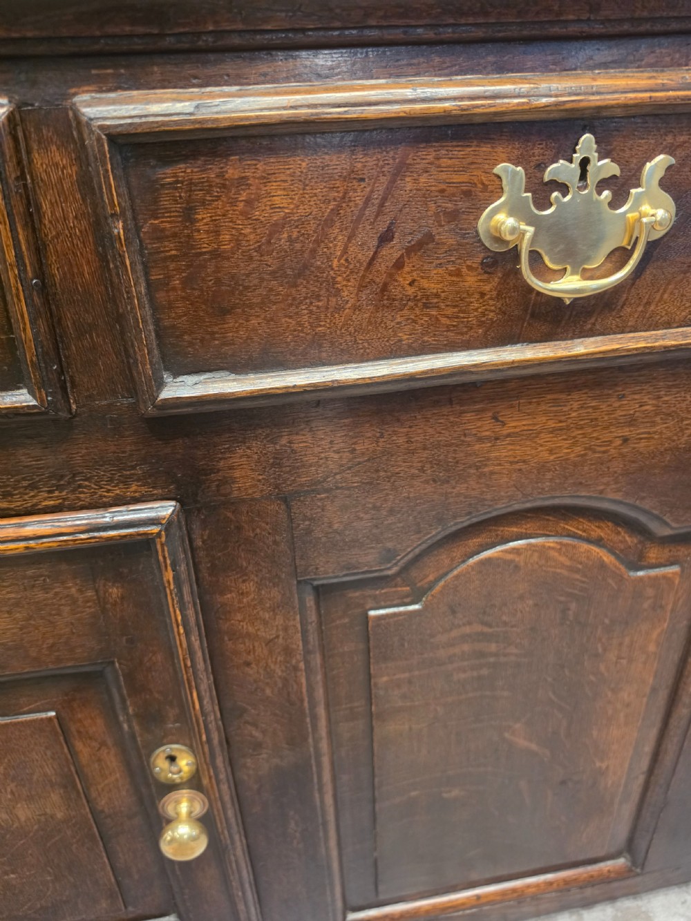An 18th-century dark oak Welsh dresser featuring a three-tiered plate rack top above three drawers and three panelled cupboard doors with brass handles. View V
