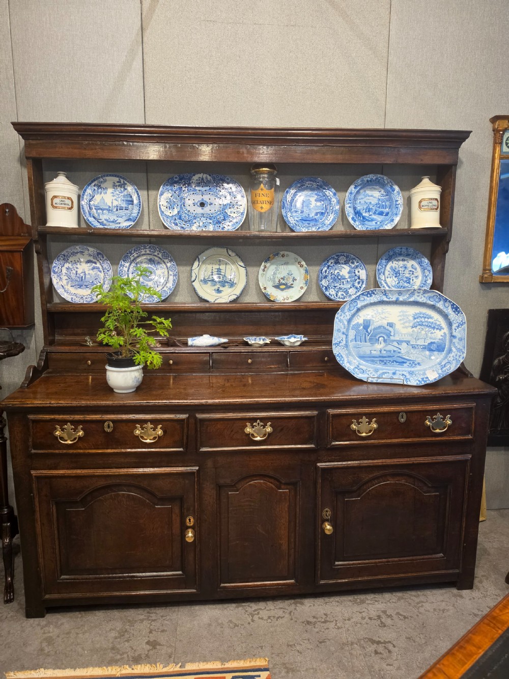 An 18th-century dark oak Welsh dresser featuring a three-tiered plate rack top above three drawers and three panelled cupboard doors with brass handles. View VIII