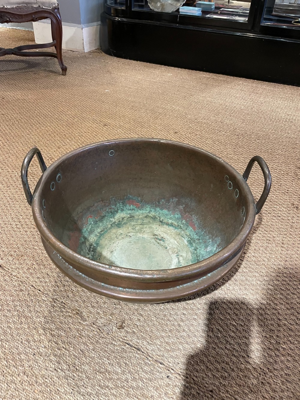 A round, hand-hammered antique copper pot with dual iron side handles and a weathered green patina on the interior base. View VI