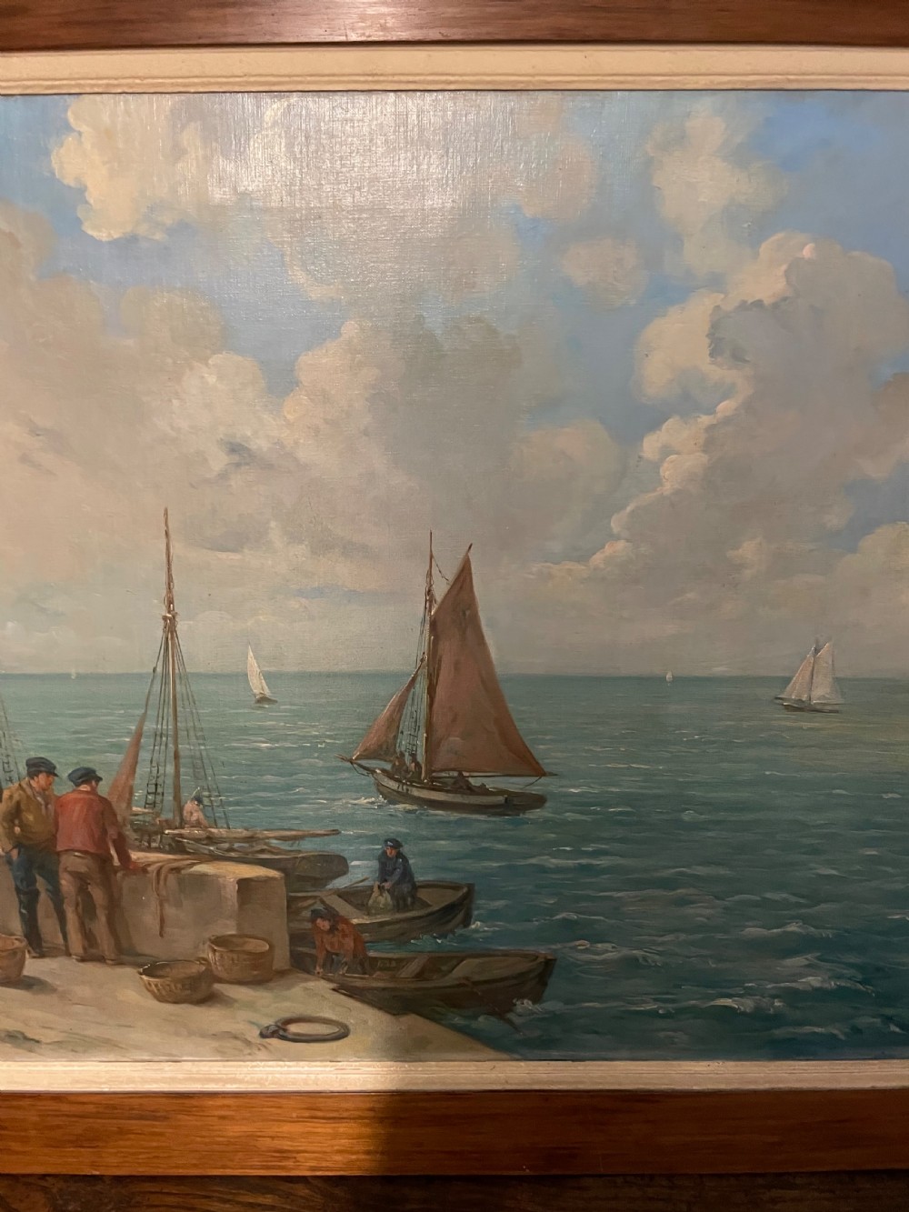 An early 20th-century oil painting on canvas depicting fishermen and sailing vessels at a stone harbour quay, set within a wide wooden frame. View III