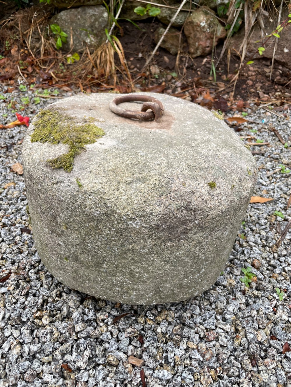 A cylindrical grey granite block rests on gravel, featuring an embedded rusted iron ring and patches of green moss on the surface.