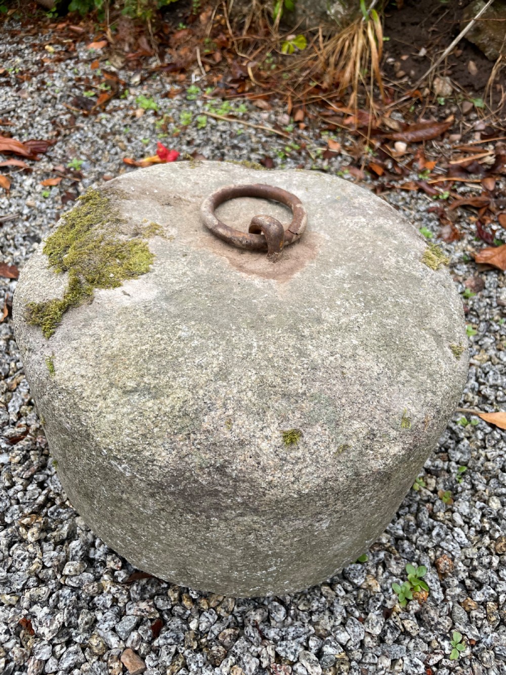 A cylindrical grey granite block rests on gravel, featuring an embedded rusted iron ring and patches of green moss on the surface. View II