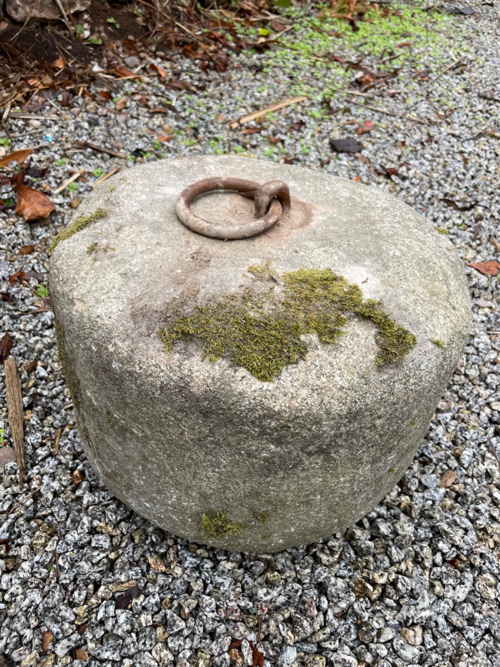 A cylindrical grey granite block rests on gravel, featuring an embedded rusted iron ring and patches of green moss on the surface. View III
