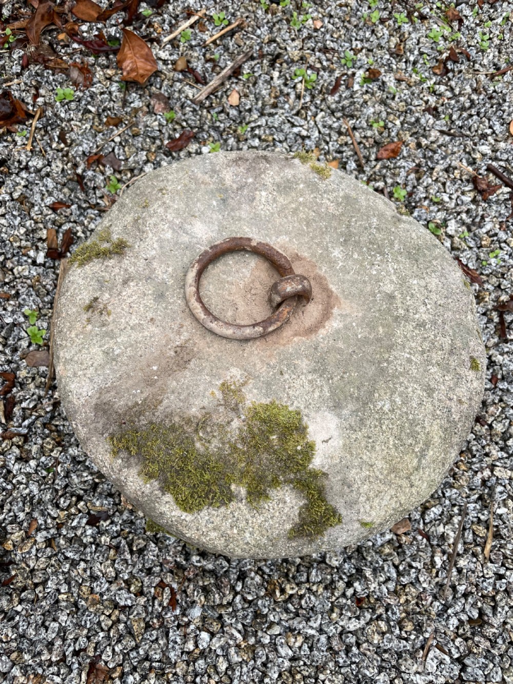 A cylindrical grey granite block rests on gravel, featuring an embedded rusted iron ring and patches of green moss on the surface. View IV