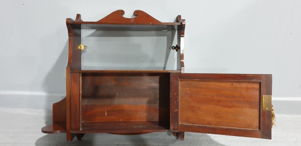 A wall-mounted warm brown mahogany cabinet featuring a single fielded panel door with a key tassel, an upper shelf with a carved scroll pediment, and lower side shelf brackets. View IV