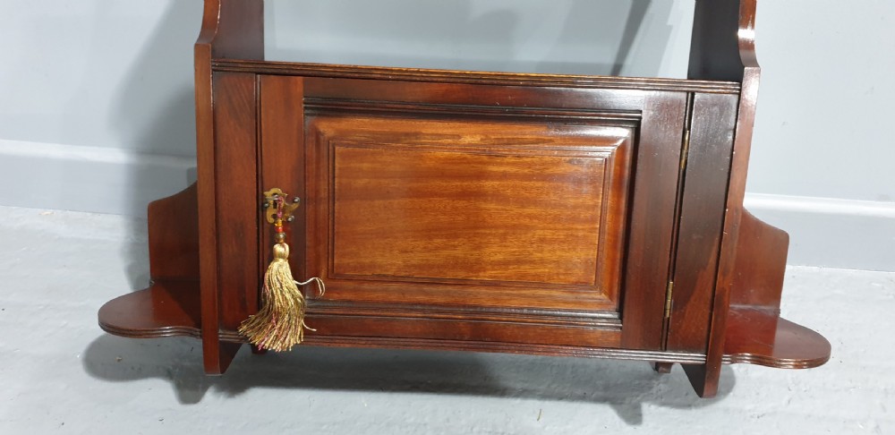 A wall-mounted warm brown mahogany cabinet featuring a single fielded panel door with a key tassel, an upper shelf with a carved scroll pediment, and lower side shelf brackets. View V