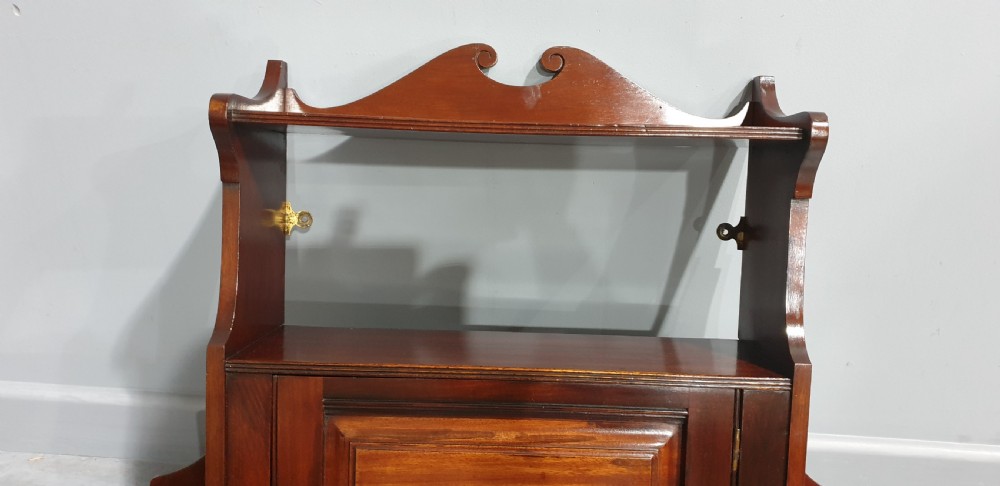 A wall-mounted warm brown mahogany cabinet featuring a single fielded panel door with a key tassel, an upper shelf with a carved scroll pediment, and lower side shelf brackets. View VI
