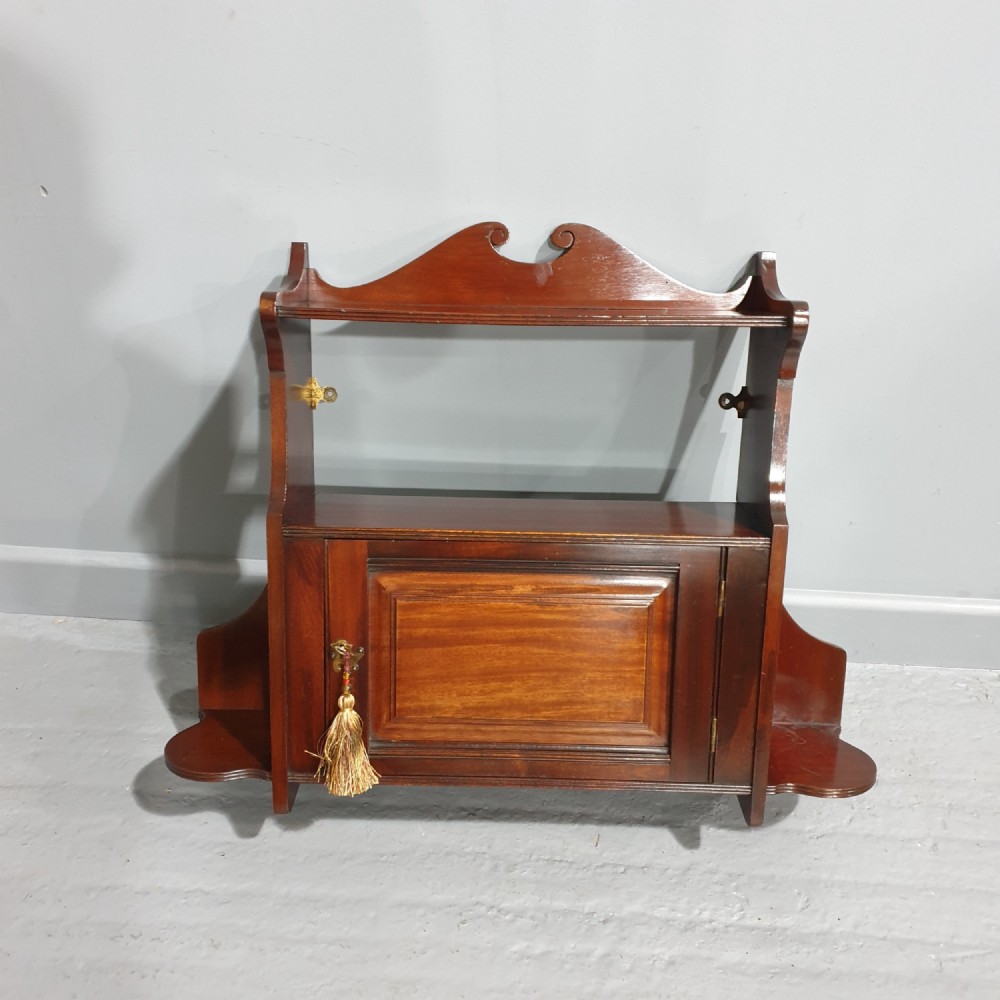 A wall-mounted warm brown mahogany cabinet featuring a single fielded panel door with a key tassel, an upper shelf with a carved scroll pediment, and lower side shelf brackets.