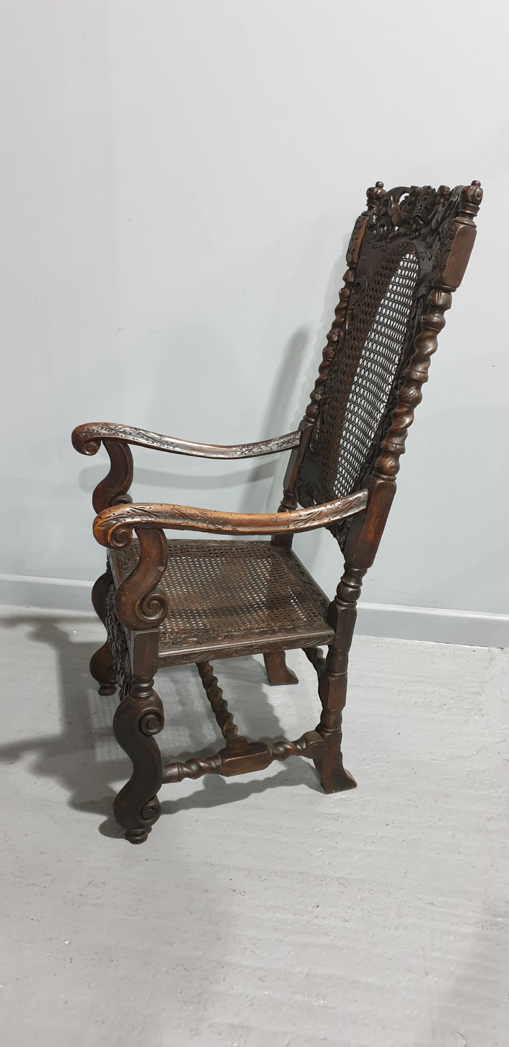A high-backed dark oak carver chair featuring an oval cane backrest, barley twist posts, hand-carved cresting, and a patterned seat cushion. View X