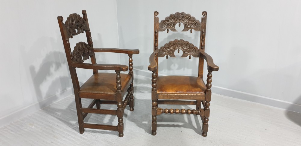 A pair of dark carved oak carver armchairs featuring decorative scrolled backrests, turned legs, bobbin stretchers, and leather seats. View V