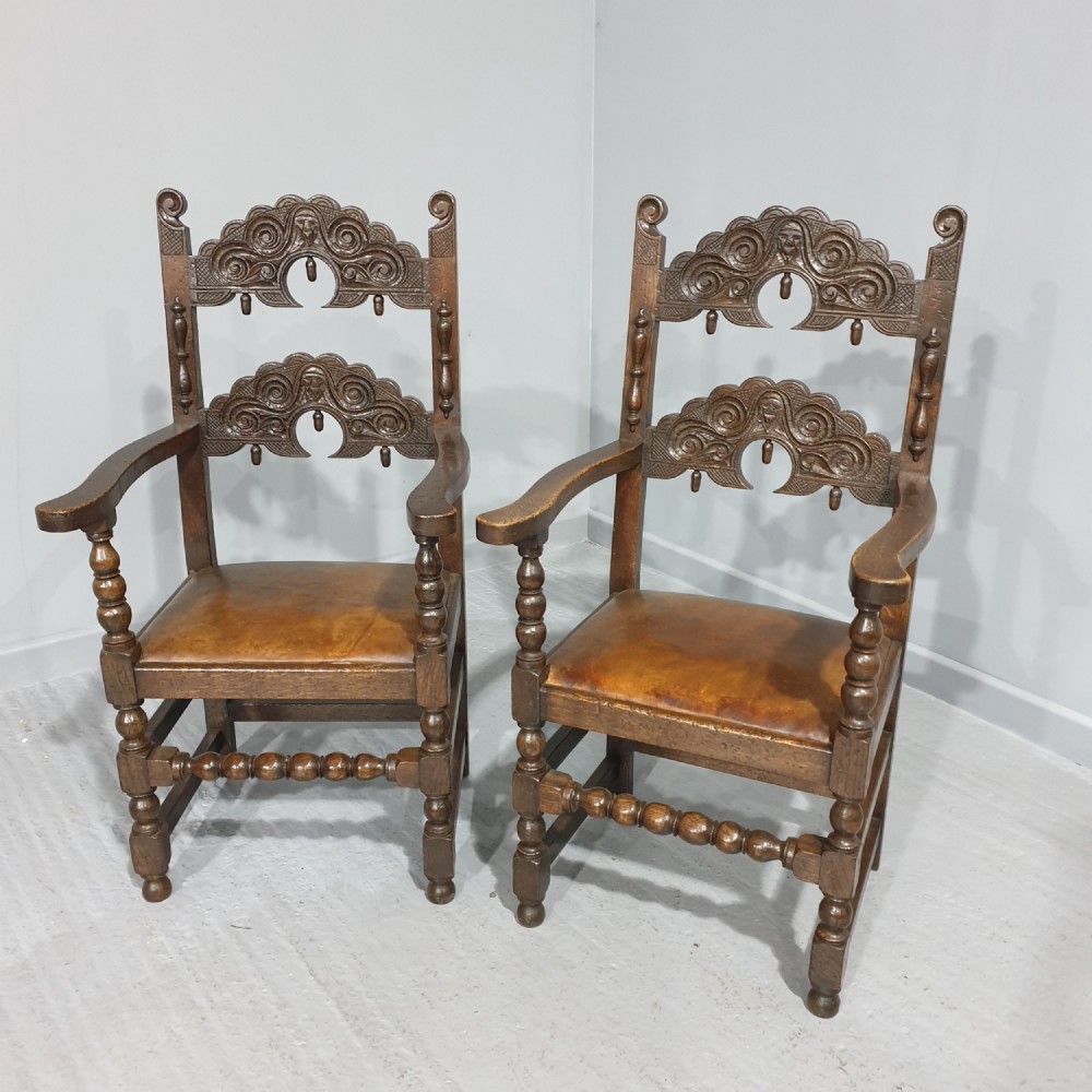 A pair of dark carved oak carver armchairs featuring decorative scrolled backrests, turned legs, bobbin stretchers, and leather seats.