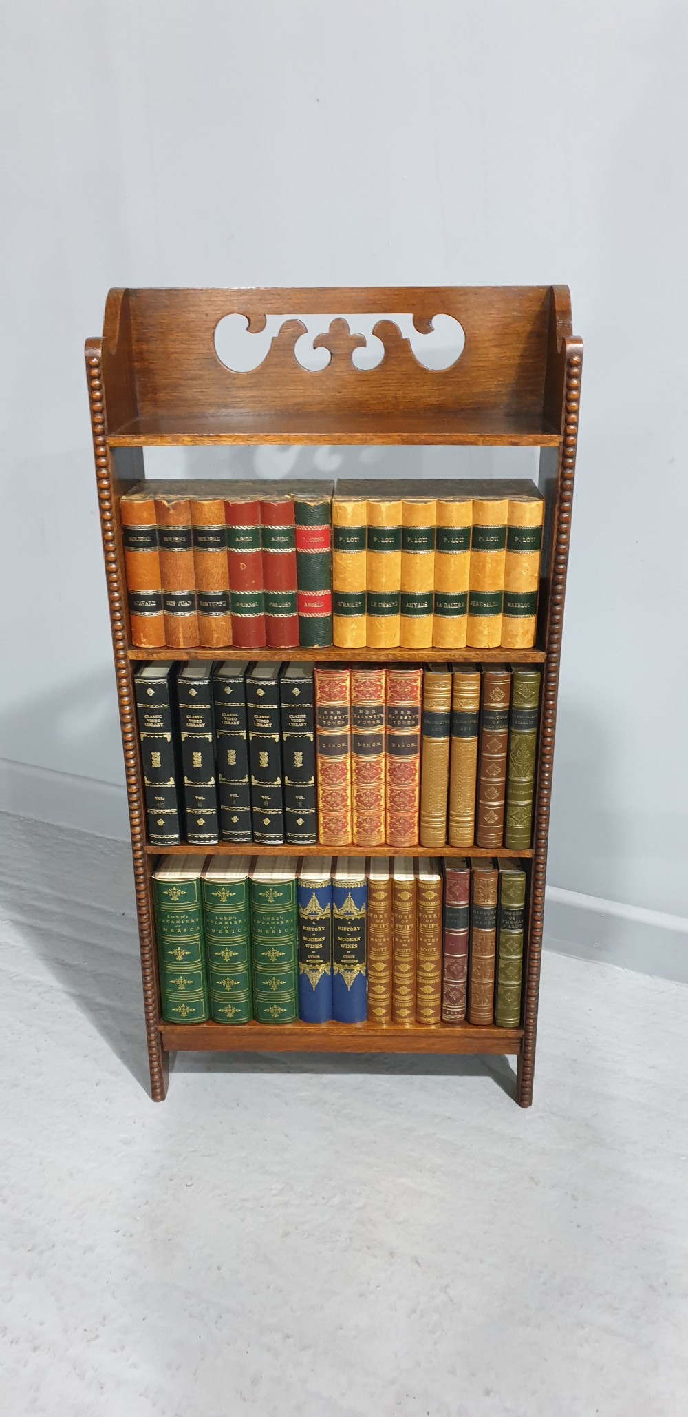 A slim freestanding open oak bookcase with three shelves, bobbin turned edge moulding, and a carved cresting top gallery rail. View IV