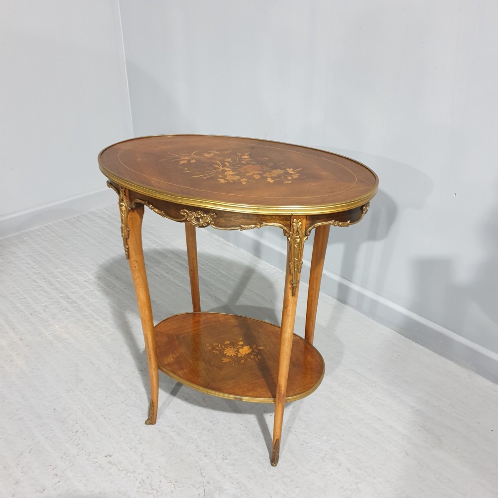 An oval French-style two-tier occasional table featuring floral marquetry inlay, slender cabriole legs, and gilt-bronze mounts.