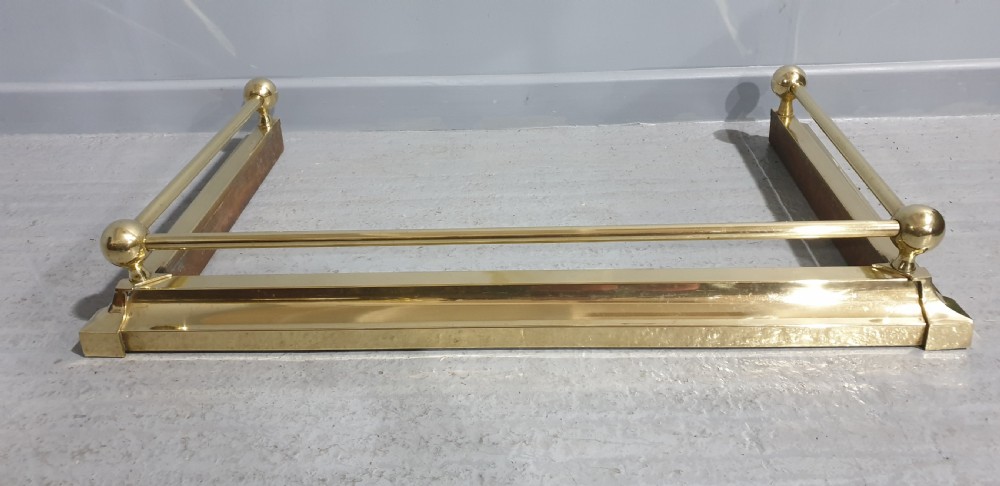 A polished Victorian brass fireplace fender featuring tubular upper rails and spherical corner finials set on a stepped base.