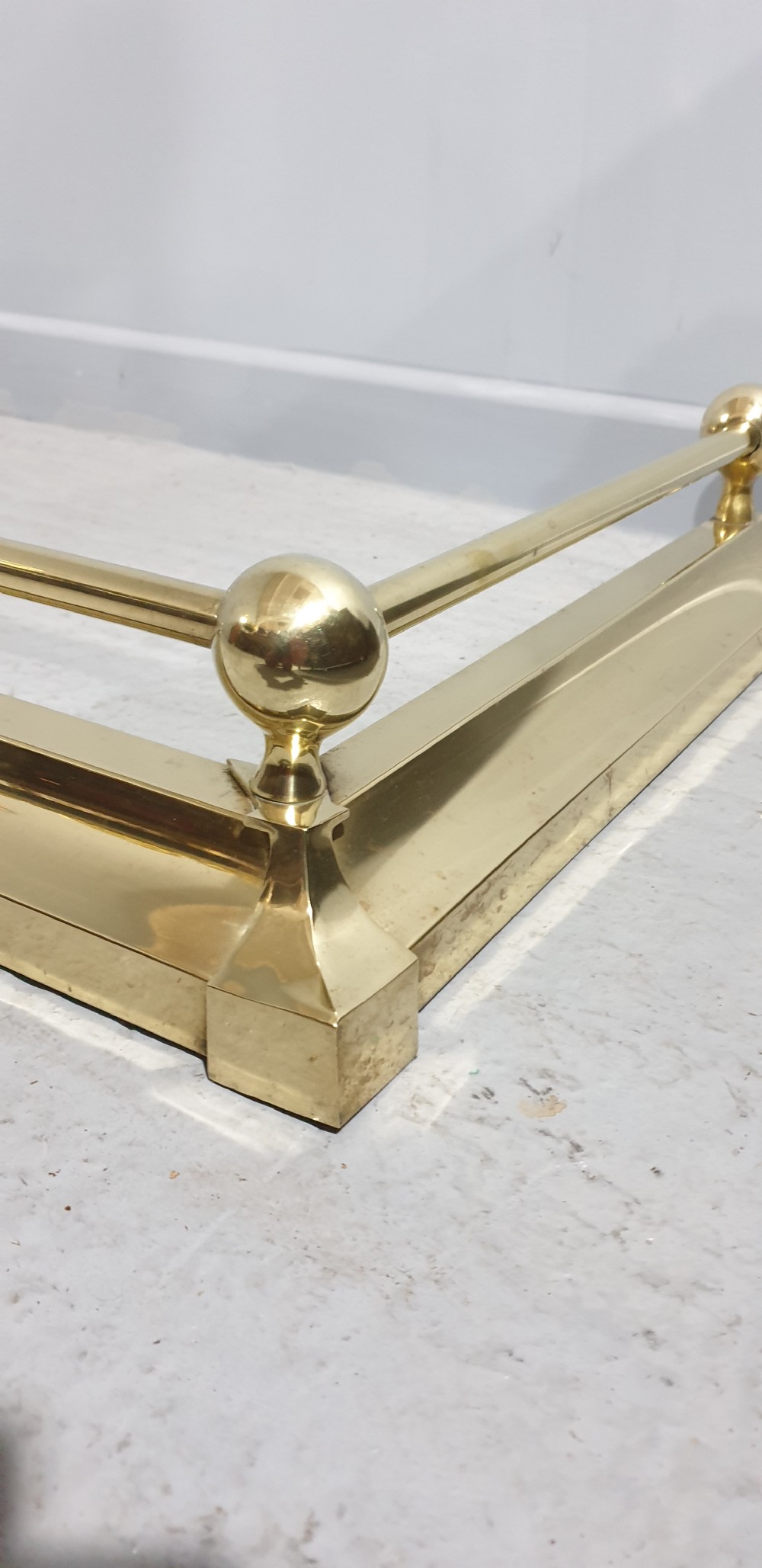 A polished Victorian brass fireplace fender featuring tubular upper rails and spherical corner finials set on a stepped base. View IV