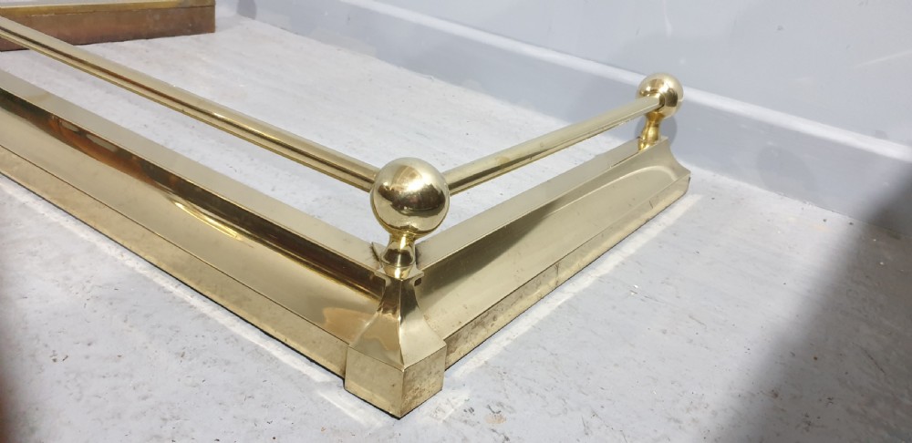 A polished Victorian brass fireplace fender featuring tubular upper rails and spherical corner finials set on a stepped base. View V